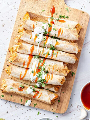 Wooden board with baked chicken taquitos with dipping sauce. White marbled surface.