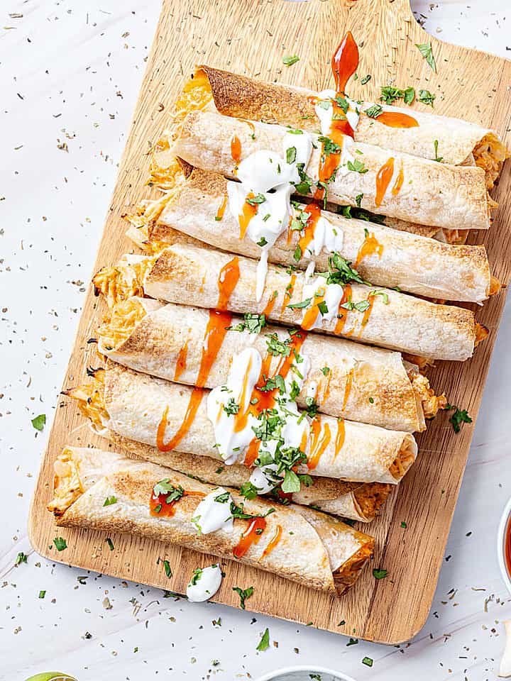 Wooden board with baked chicken taquitos with dipping sauce. White marbled surface.
