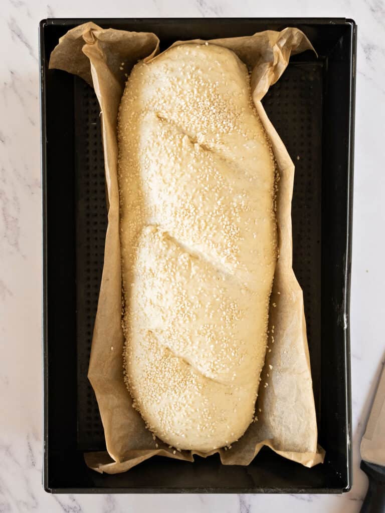 Scored, sesame-crusted bread loaf before baking on parchment paper and black tray.