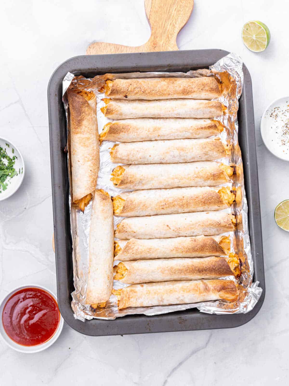 Oven tray with baked taquitos. White marbled surface, bowls with garnishes.