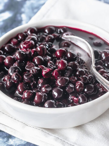 Quick blueberry compote in white bowl with silver spoon. Blue white cloth surface.