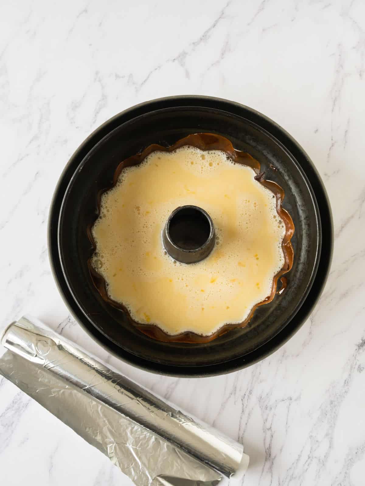 Bundt cake pan with chocoflan mixture. White marbled surface. Roll of foil.