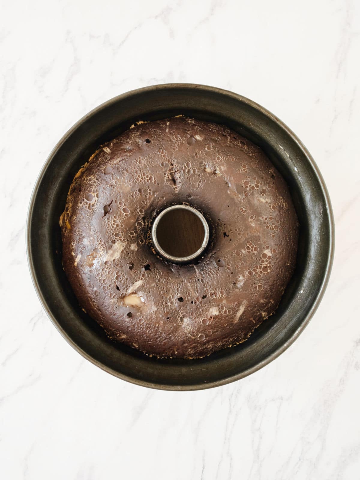 Baked chocolate flan cake in a dark bundt pan. White marbled surface.