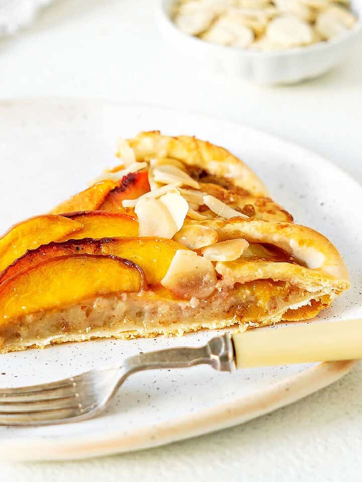 Almond cream peach tart slice on a white plate. Silver fork. White background.