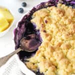 Partial view of blueberry pineapple dump cake. White round dish, white surface. Silver spoon.