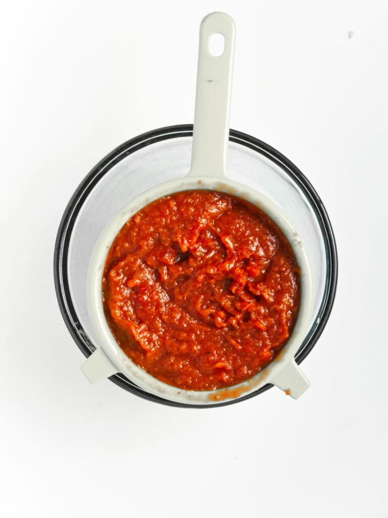 Draining homemade ketchup over a glass bowl. White surface.