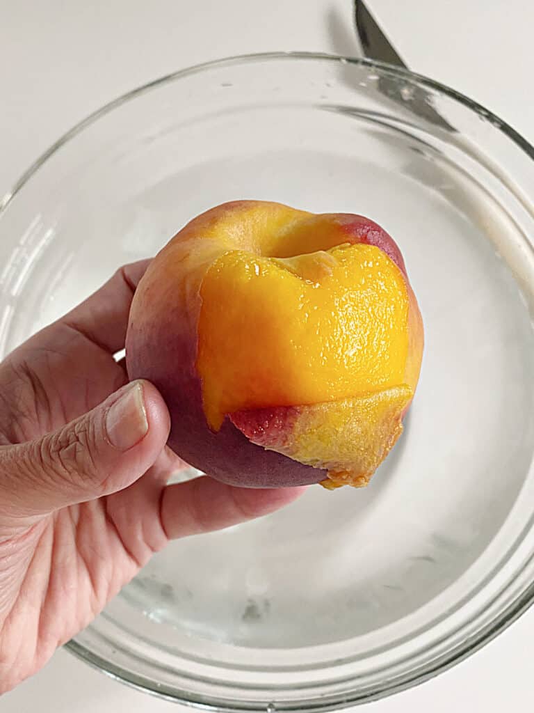 Half peeled fresh peach over a glass bowl with water.