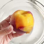 Close up half peeled fresh peach over a glass bowl with water.