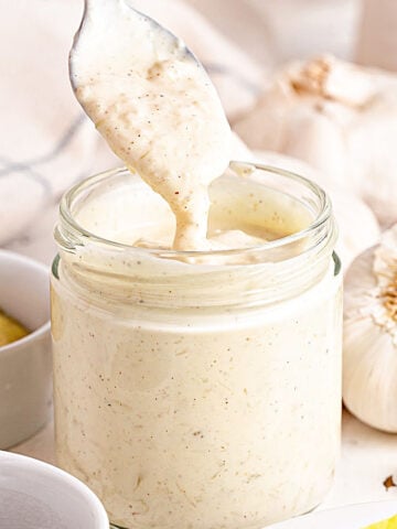 Jar with garlic aioli being lifted with a spoon. White bowls, garlic heads, white background.