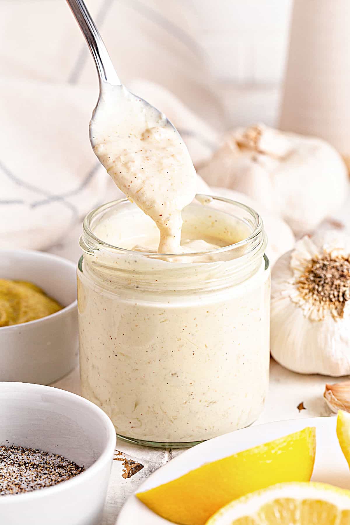 Silver spoon lifting aioli from a jar. White background with garlic cloves, lemon wedges and bowls. 