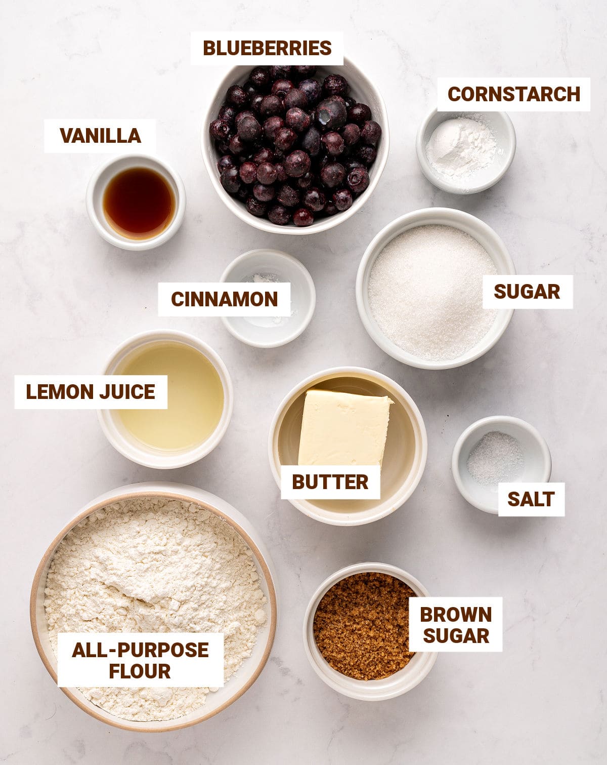 Ingredients for blueberry crumble in bowls on a white surface including lemon, blueberries, butter, vanilla and sugars.