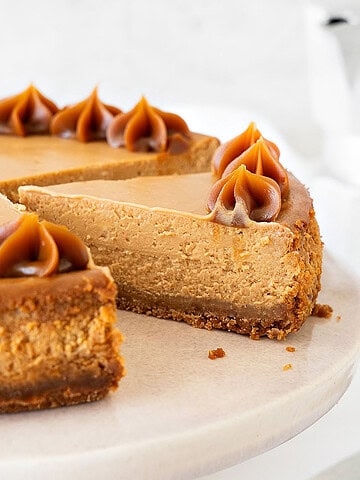 Whole and slice of dulce de leche cheesecake on a pink cake stand. White background.