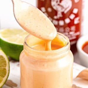 Close up jar and spoon with spicy aioli. Limes and sriracha bottle in the background.