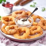 Beige platter with onion rings around a bowl with dip. Spoon, grayish cloth.