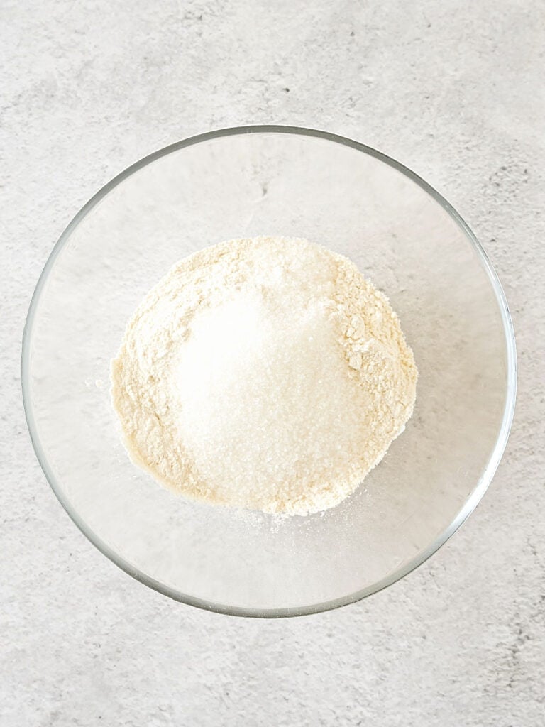 Flour mixture and sugar in a glass bowl on a light gray surface. 