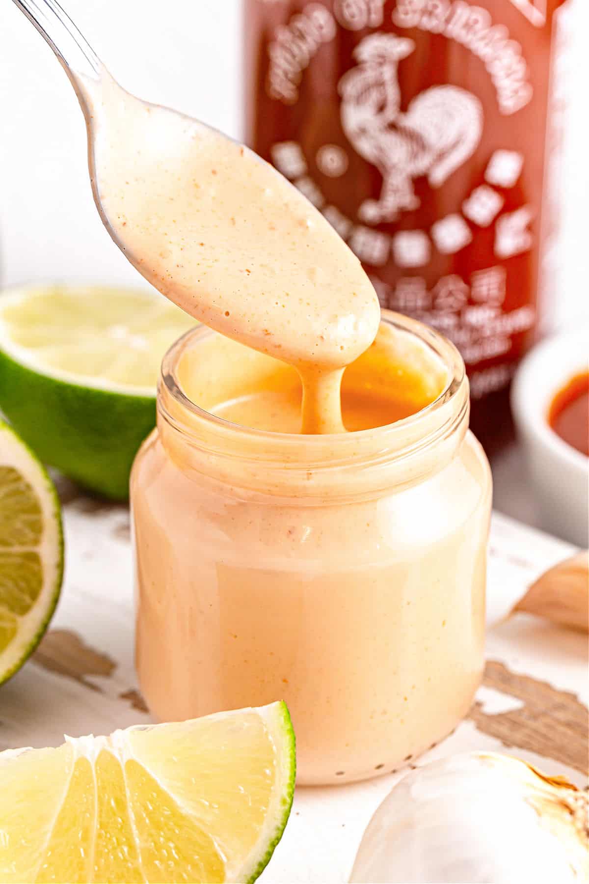 Spooning spicy aioli from a jar. Lime wedges, white surface, sriracha bottle.