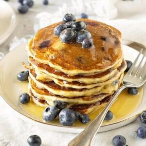 Several stacked ricotta blueberry pancakes with syrup. White plate. Silver fork.