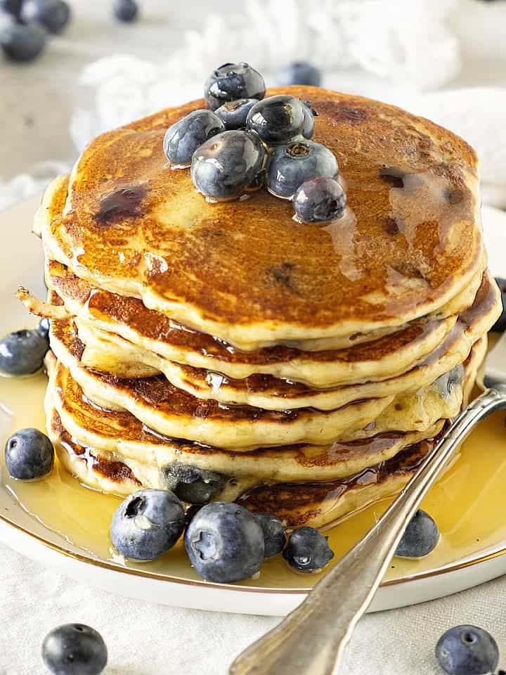 Several stacked ricotta blueberry pancakes with syrup. White plate. Silver fork.