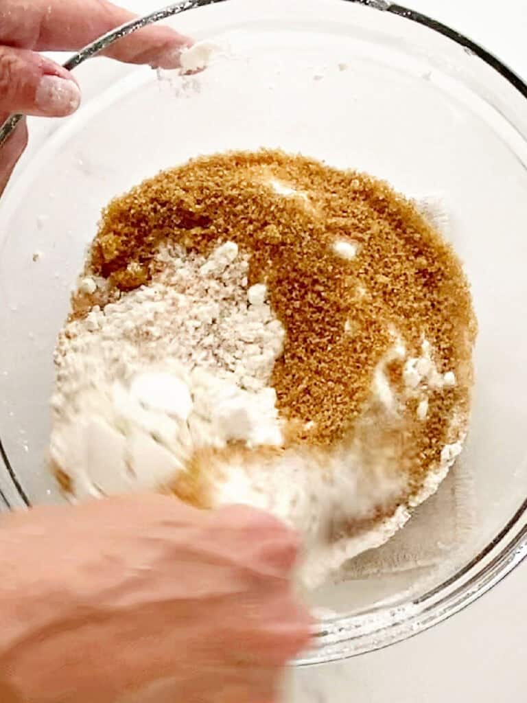 Stirring brown sugar into flour mixture in a glass bowl. White background.