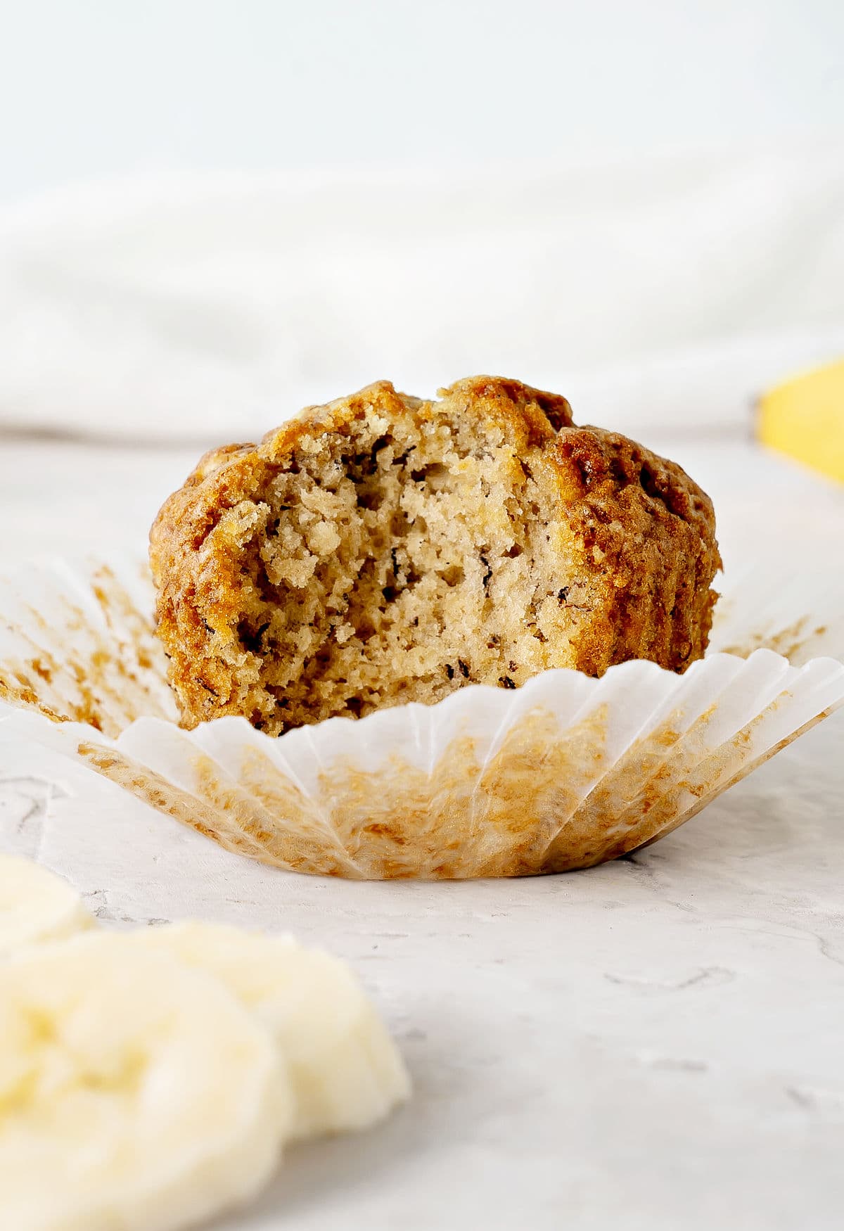 Open paper liner with bitten banana muffin. Light gray background. Banana slices.