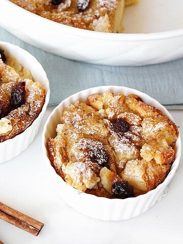 Two white bowls with raisin bread pudding. White and blue surface, cinnamon sticks.