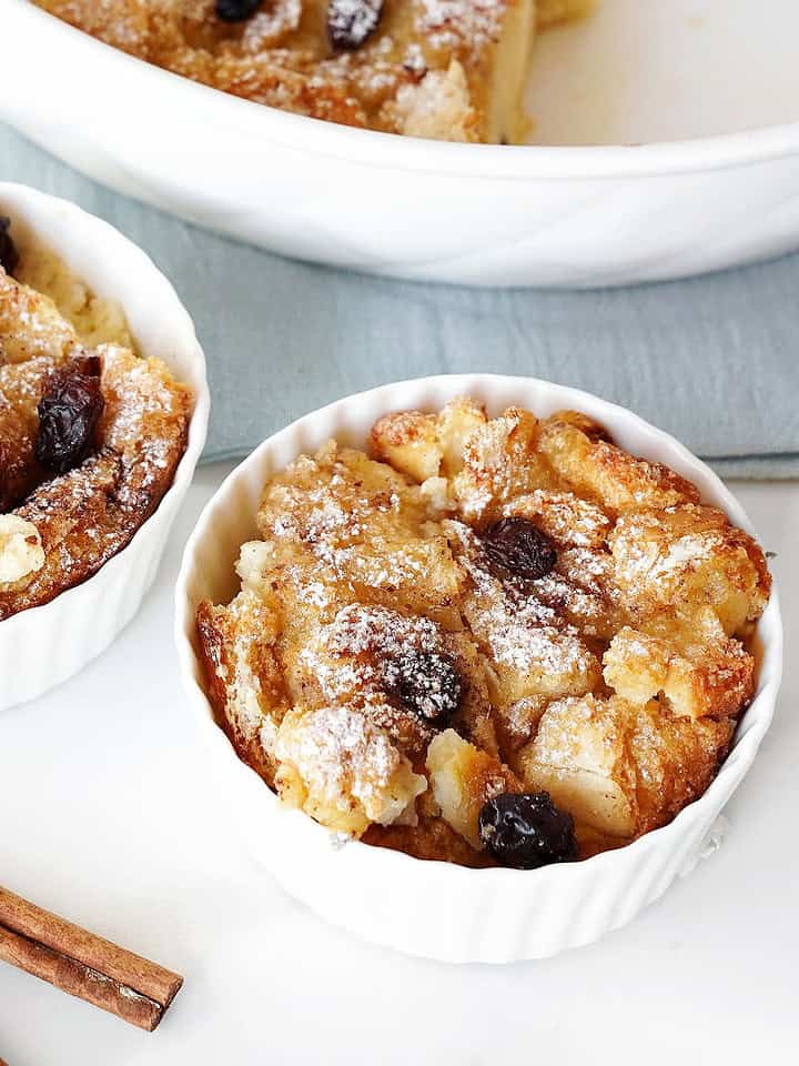 Two white bowls with raisin bread pudding. White and blue surface, cinnamon sticks.