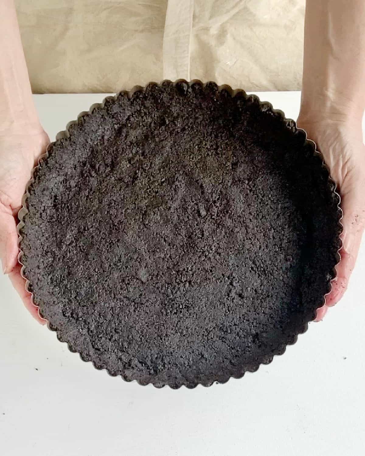 Hands holding chocolate cookie crust over a white and beige background. 