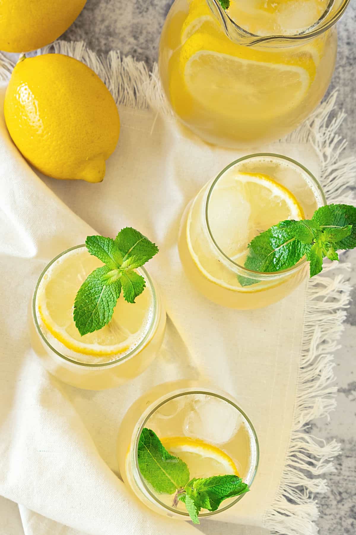 Pitcher and glasses of ginger lemonade with mint sprigs. White cloth and whole lemons.