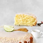Glazed slice of coconut cake being lifted from whole cake. Light gray background.