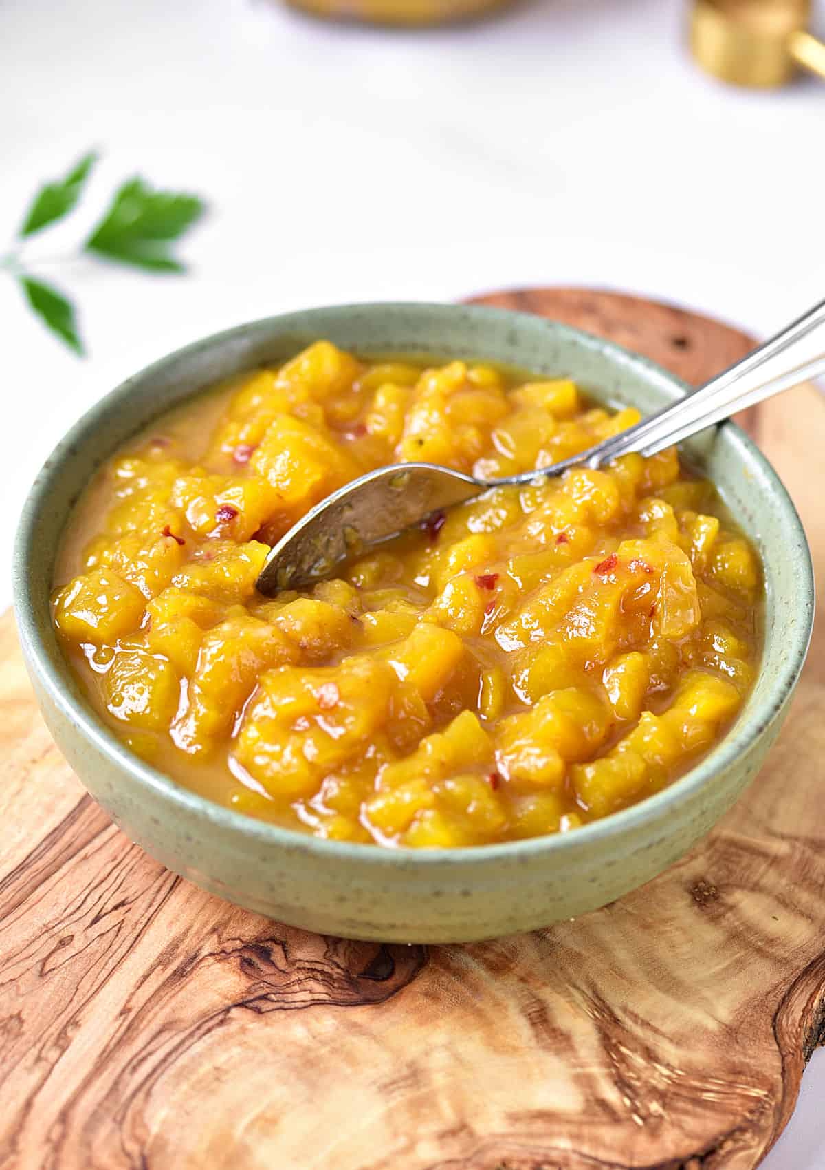 Wooden board with green bowl containing mango chutney. A silver spoon, white background.