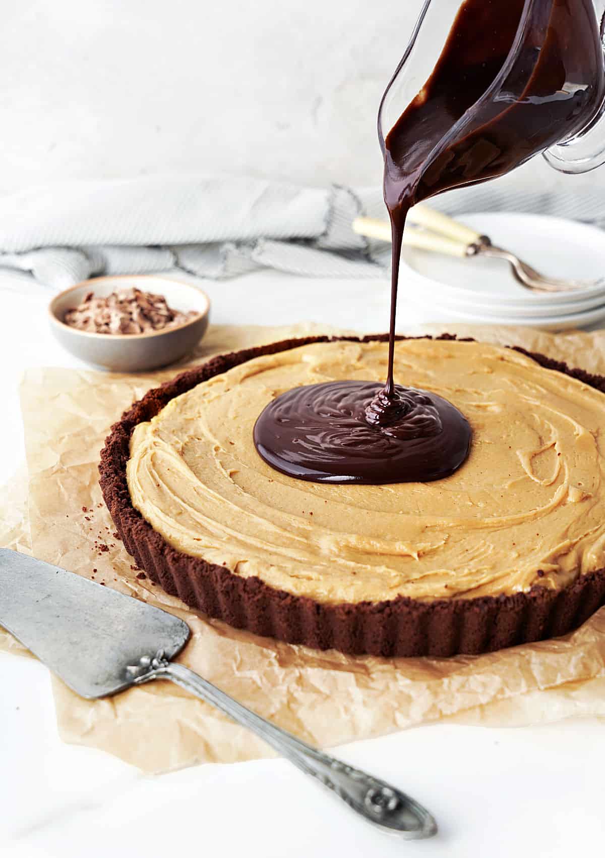 Pouring chocolate ganache sauce on peanut butter pie on a beige parchment paper. White background, silver cake server.