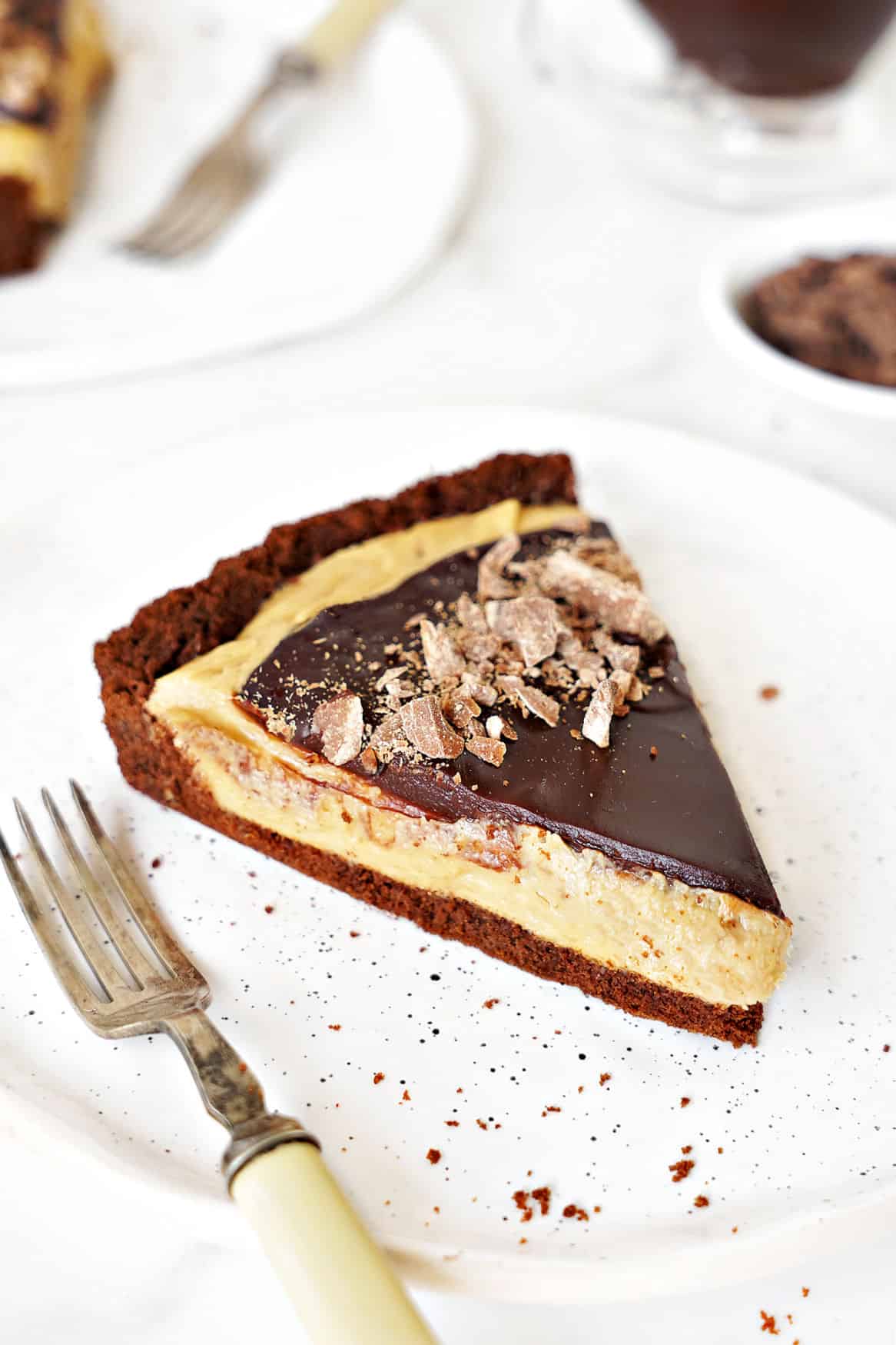 Slice of chocolate peanut butter tart on a white plate. White surface, bowl with chocolate, sauce boat. 