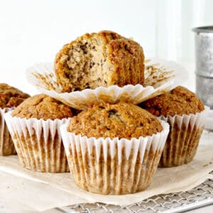 Bitten banana muffin on top of whole ones in paper liners. Beige and white background.