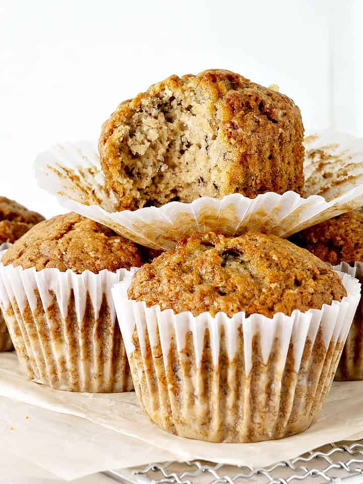 Bitten banana muffin on top of whole ones in paper liners. Beige and white background.