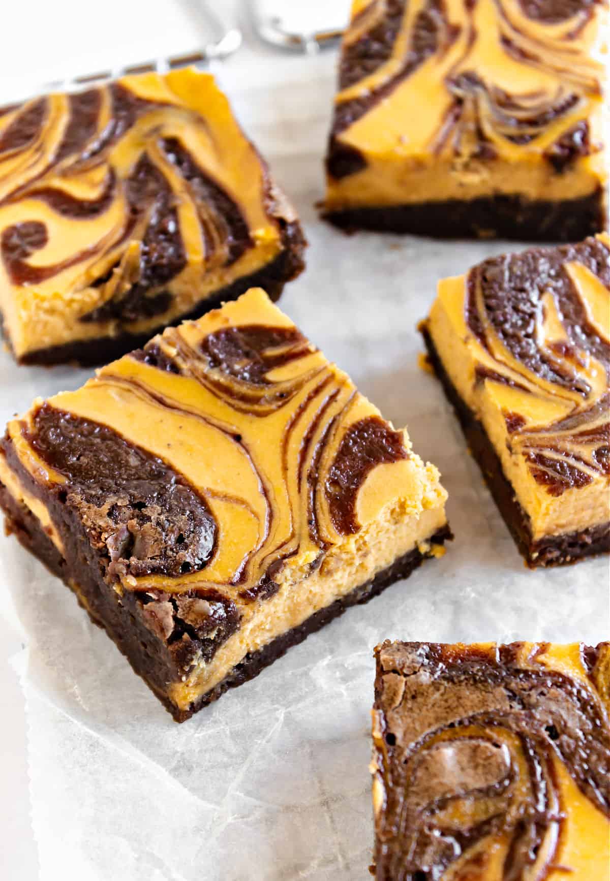 Several squares of brownies with pumpkin cheesecake swirl on white parchment paper. 