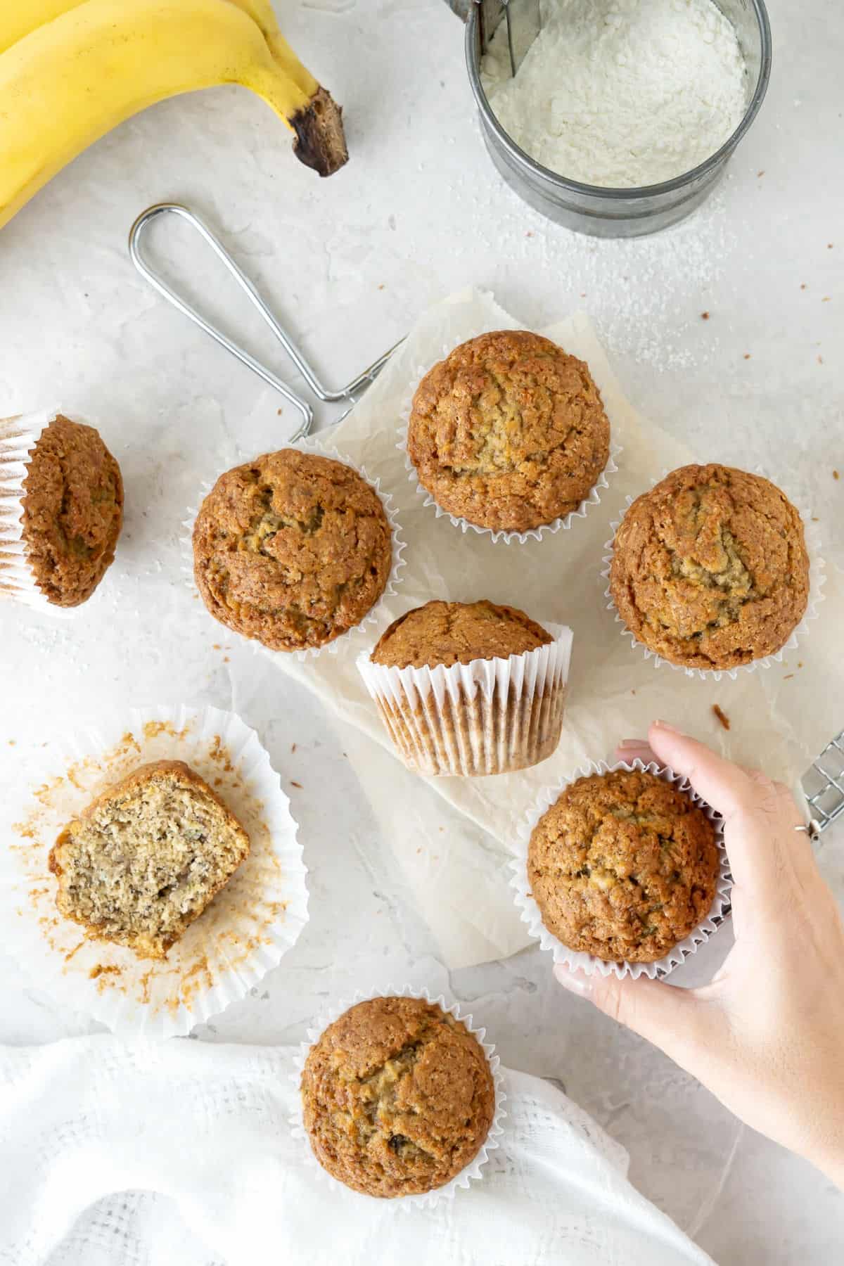 Top view of several banana muffins on a light gray surface. A hand, a banana, white cloth.