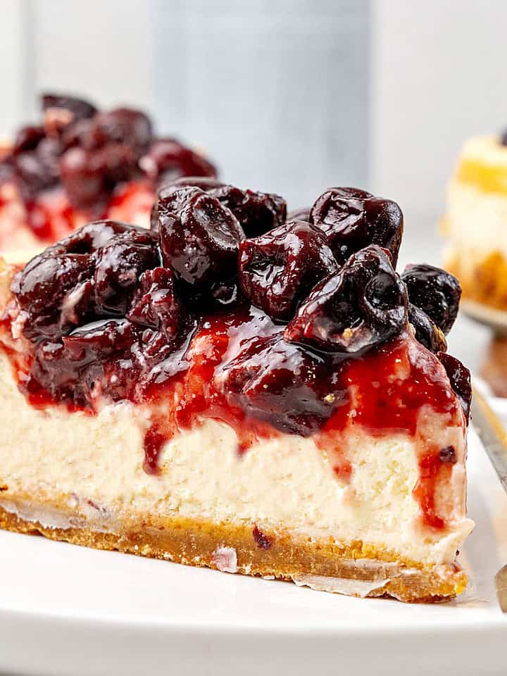 Close up slice of cherry-topped cheesecake. White plate, gray background.