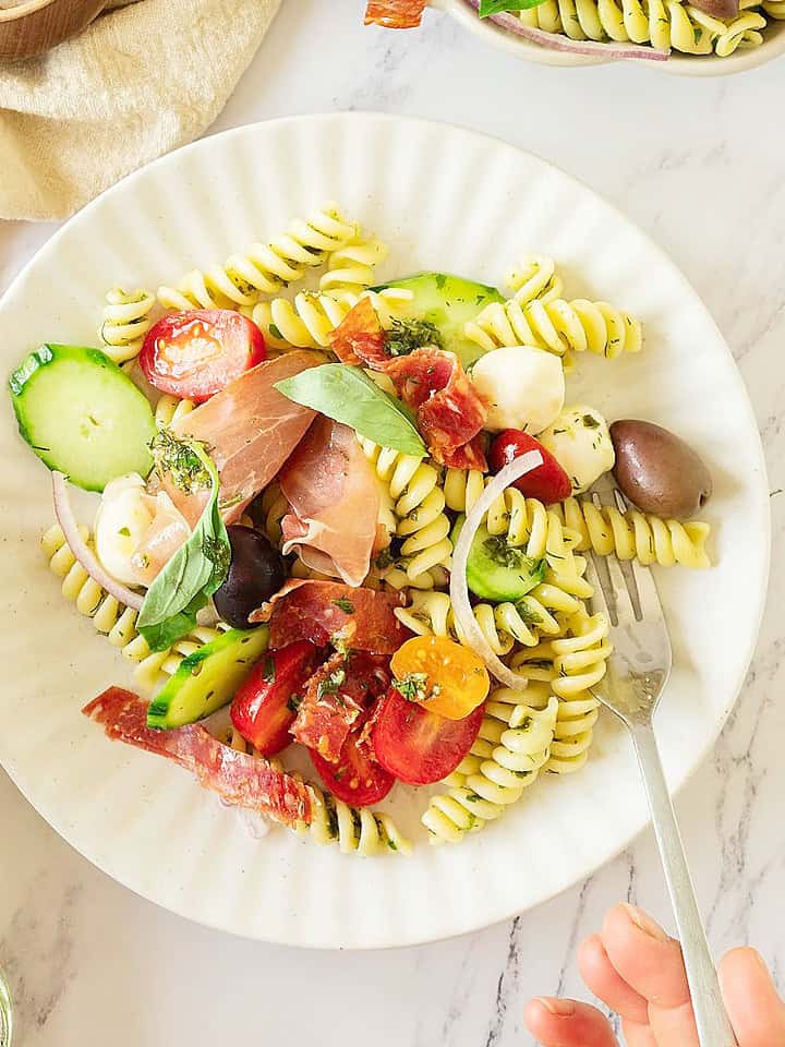 Hands holding silver fork and white plate with antipasto pasta salad. White surface.