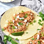 Paper-lined metal tray with shredded chicken tacos. Lime wedges, cilantro.