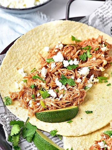 Paper-lined metal tray with shredded chicken tacos. Lime wedges, cilantro.