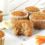 Wire rack with bitten and whole carrot banana muffins. White background. Banana slices, carrot sticks.
