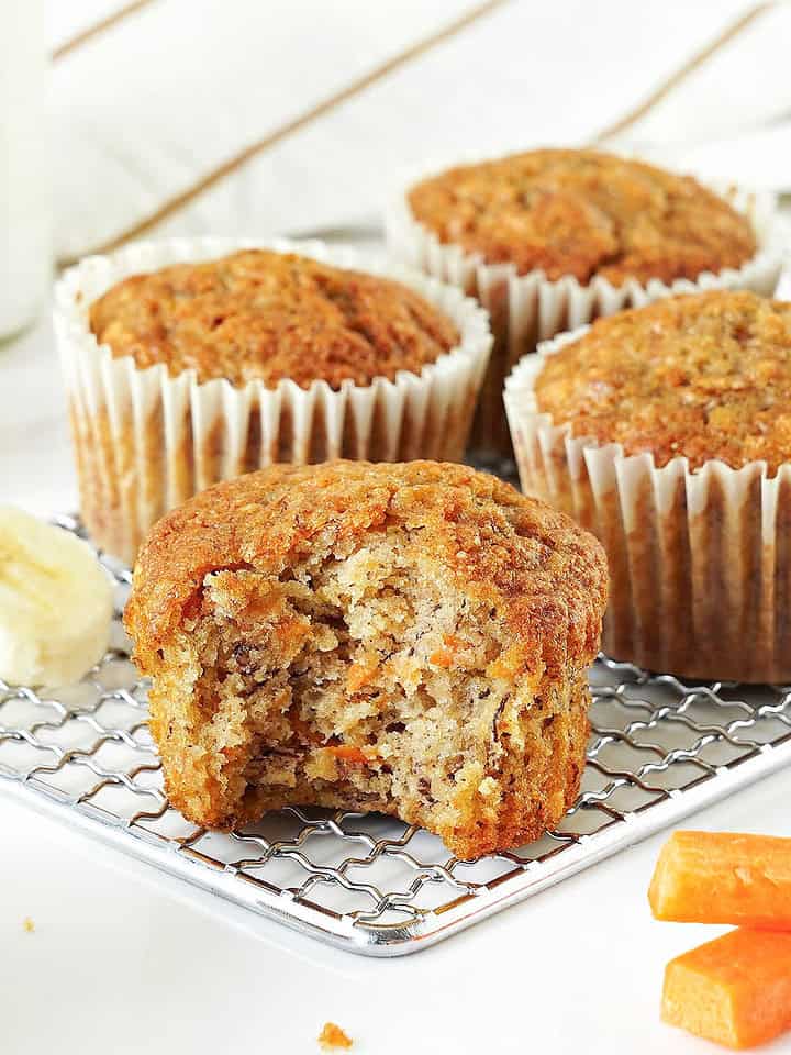 Wire rack with bitten and whole carrot banana muffins. White background. Banana slices, carrot sticks.