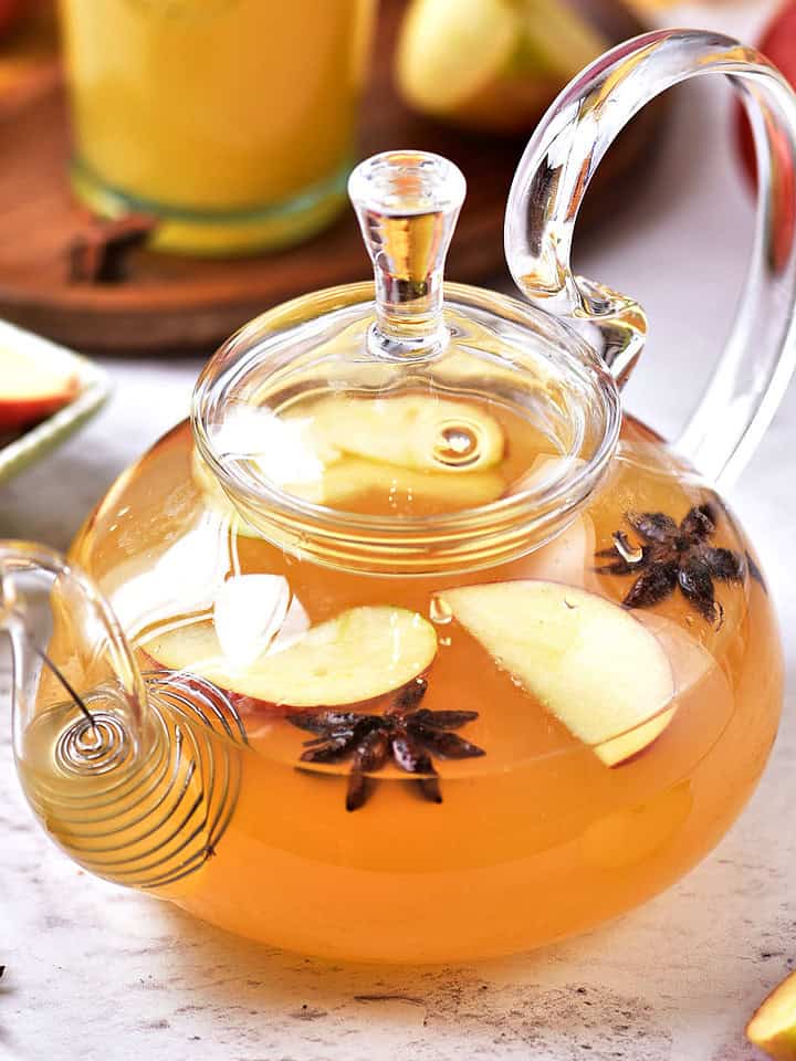 White gray surface with spiced apple cider in a glass teapot. Apples and glass in the background.