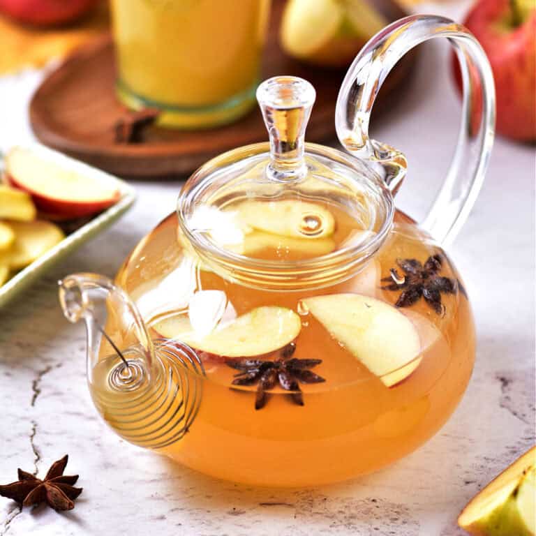 White gray surface with spiced apple cider in a glass teapot. Apples and glass in the background.