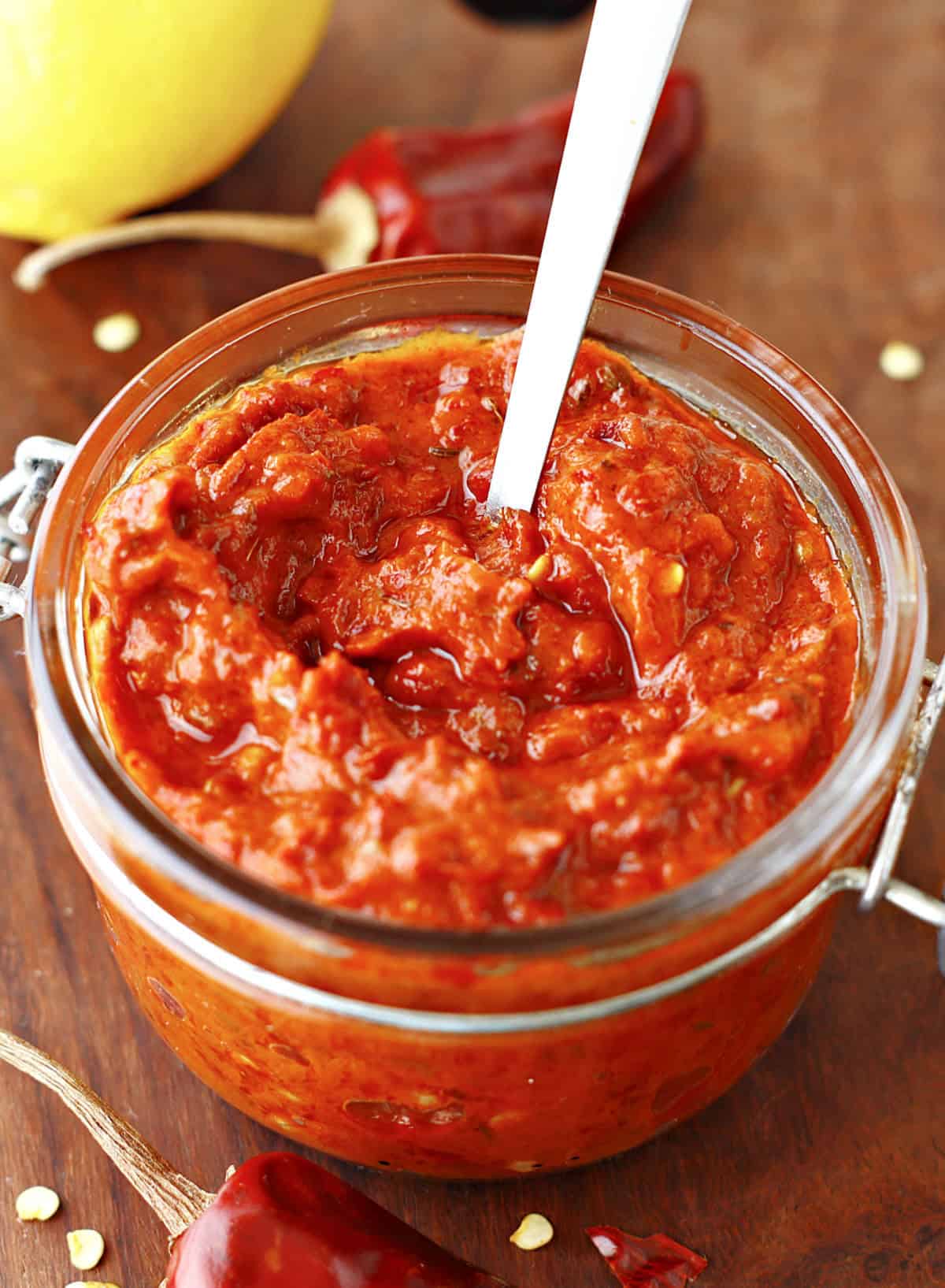 Harissa paste in a mason jar with silver spoon inside. Wooden board, dried chiles around.