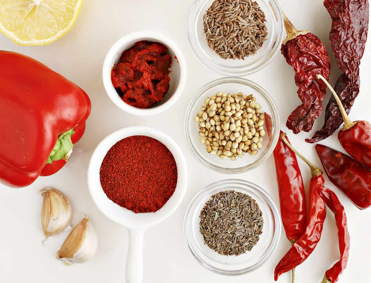 White bowls and surface with ingredients for harissa including dried chiles, garlic, spices and lemon.