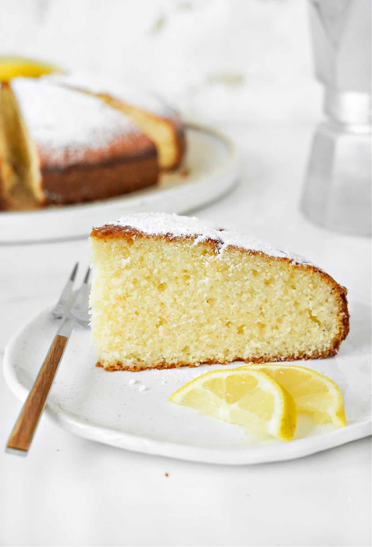 Single slice of powdered sugar-topped ricotta cake. White plate, a fork, lemon slices. Whole cake in white background.