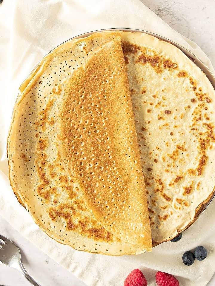 Whole thin pancakes on a plate. White towel and surface. Whole berries around. Top view.