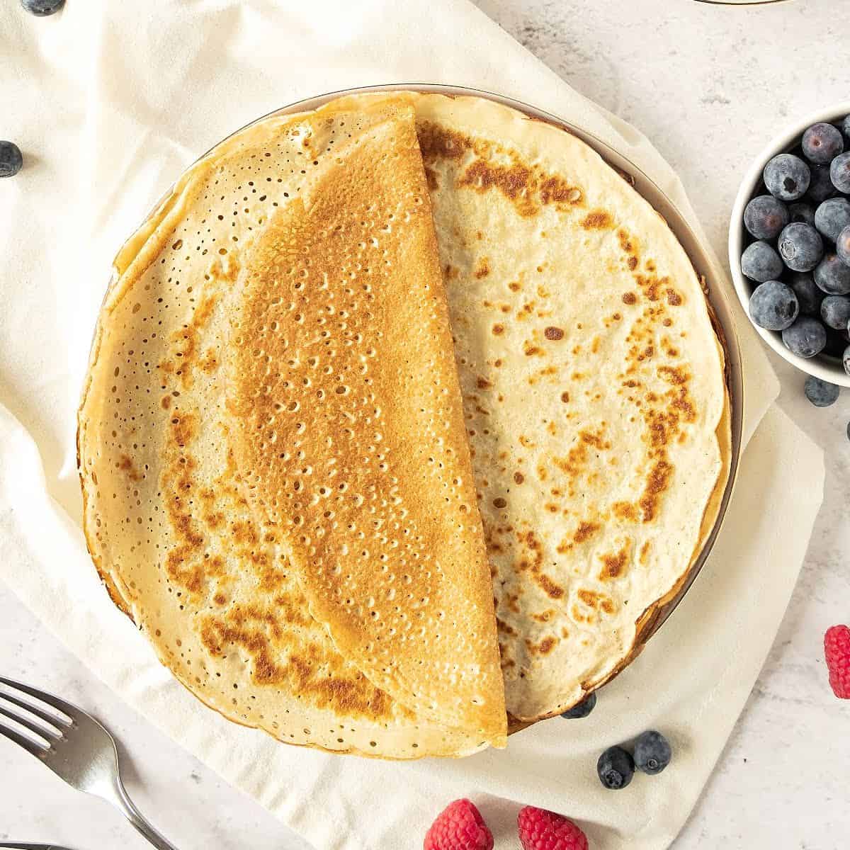 Whole thin pancakes on a plate. White towel and surface. Whole berries around. Top view.