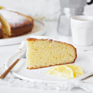 White plate with ricotta cake slice. A fork, lemon slices, white background with whole cake.